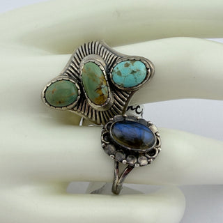 A pair of silver rings displayed on a white mannequin hand. The larger ring features three oval turquoise-colored stones with brown matrix details, and the smaller ring features an oval dark blue stone surrounded by small silver dots.