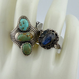 Two silver rings displayed on a white ceramic ring stand. The left ring features three turquoise-colored stones in a textured silver setting, and the right ring features a blue oval stone surrounded by dark round stones in an ornate silver setting.