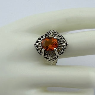 An ornate silver ring with a large oval orange gemstone, displayed on a white ceramic ring stand against a light gray background.