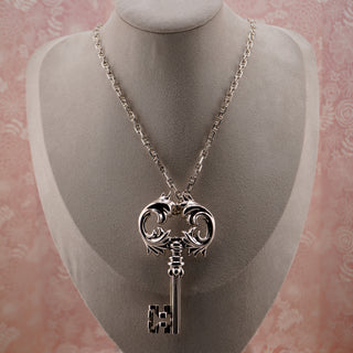 Silver key-shaped pendant on a chain displayed on a mannequin bust against a pink floral patterned background.