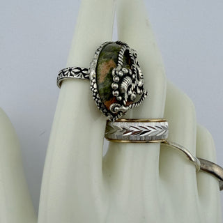 A silver ring with a large oval green and brown stone centerpiece, displayed on a white ring stand alongside another silver ring with a chevron pattern.