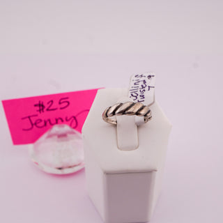 A twisted rope-style ring with a silver-toned band and dark oxidized detailing, displayed on a white jewelry stand. A pink handwritten price tag reading '$25 Jenny' and a small clear glass bead are visible in the background.