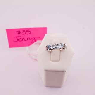 A silver band ring with five clear baguette-cut gemstones displayed on a white jewelry stand, with a pink handwritten price tag card in the background.