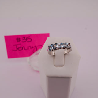 A silver band ring with a row of rectangular light blue gemstones, displayed on a white jewelry stand. A pink handwritten price tag card is visible in the background on a pale pink surface.