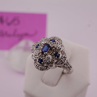 An ornate silver filigree ring with a central rectangular blue gemstone and small blue accent stones, displayed on a white ring stand. A pink card with cursive writing is blurred in the background.