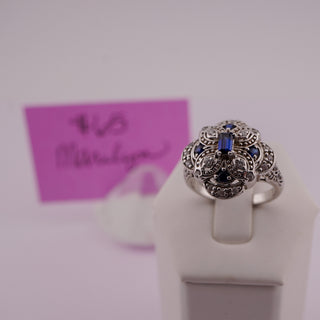 An ornate silver filigree ring with a central blue rectangular gemstone and small blue and black stones, displayed on a white stand against a light background with a pink handwritten price card in the background.
