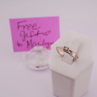A silver ring with a small rectangular gold accent on the band, displayed on a white ring stand. A pink handwritten card reading 'Free gifts to Marilyn' is blurred in the background.