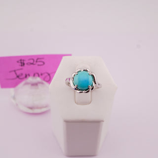 A silver ring with a round turquoise-blue gemstone displayed on a white stand, with a pink handwritten price tag in the background.