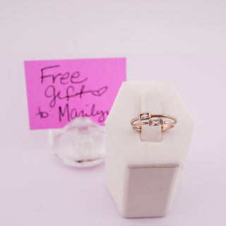 A rose gold ring with a small clear gemstone set in a white display stand, with a pink handwritten note in the background.
