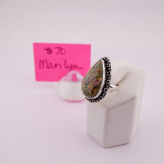A teardrop-shaped gemstone ring with a silver-toned beaded border, displayed on a white stand. A pink handwritten price card is visible in the background.