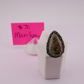A teardrop-shaped ring with a colorful iridescent stone and a black beaded metal setting, displayed on a white stand with a pink handwritten price card in the background.