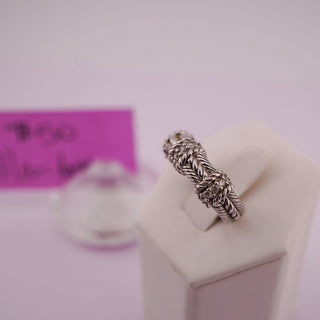 A silver-toned textured ring with small clear stones, displayed on a white jewelry stand against a pale pink background.