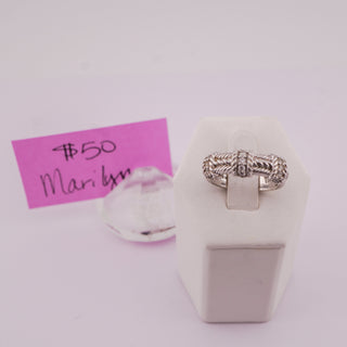 A silver-toned twisted rope ring displayed on a white jewelry stand, with a pink handwritten price tag card and a clear acrylic block in the background.