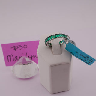 A silver ring with a row of green emerald-cut stones, displayed on a white jewelry stand with a blue tag. A pink handwritten price card is visible on the left.