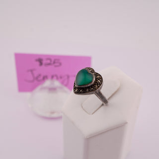 A silver ring with a heart-shaped green stone, displayed on a white stand with a pink handwritten price tag in the background.