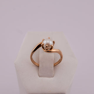 A gold ring with a single white pearl set in a curved prong setting, displayed on a white jewelry stand against a light lavender background.