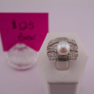 A silver ring with a central white pearl and rows of small clear stones, displayed on a white ring stand. A pink handwritten price card is blurred in the background.