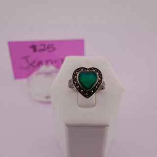 A heart-shaped ring with a green center and a dark, textured halo, displayed on a white ring stand. A pink handwritten price tag is visible in the background.