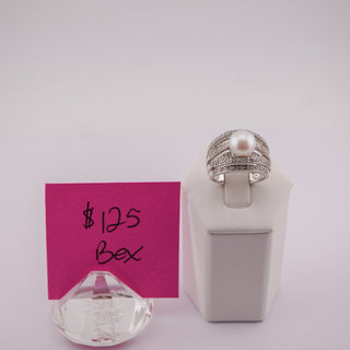 A silver ring with a central white pearl and small pavé-set stones, displayed on a white ring stand next to a pink price card reading $125 Bex, with a clear crystal prop in front.