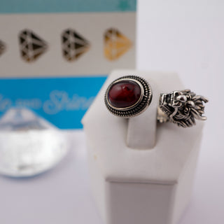 Two silver rings displayed on a white ring stand. One ring features an oval dark red cabochon stone in a textured bezel setting, and the other is a detailed silver lion head ring.