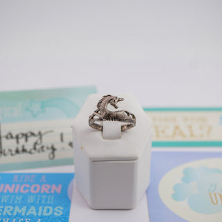 A silver ring shaped like a unicorn standing on its hind legs, displayed on a small white hexagonal stand. The background is softly blurred with pastel-colored cards.