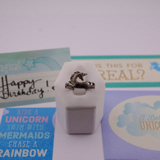 A sterling silver ring featuring a detailed unicorn design, displayed on a white geometric stand. The background is decorated with colorful unicorn-themed birthday cards.