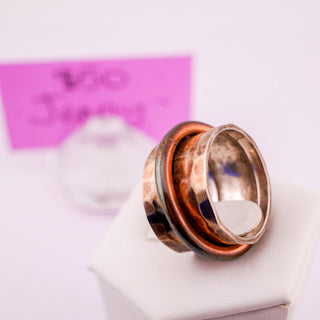 A handmade ring with stacked copper and silver bands, resting on a white display block with a pink handwritten price card in the background.