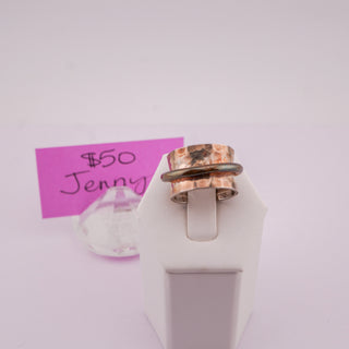 A handmade ring with textured copper and polished silver bands, displayed on a white stand with a pink handwritten price tag in the background.