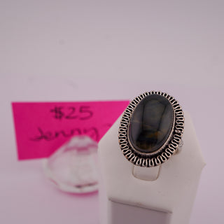 A silver oval ring with a dark, patterned stone, displayed on a white stand. A pink handwritten price tag is visible in the blurred background.