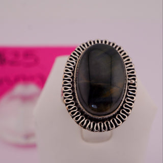 A silver ring with an oval black stone featuring subtle golden-brown veining, displayed on a white stand with a pink handwritten price tag in the background.