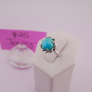 A sterling silver ring with a round turquoise cabochon gemstone, displayed on a white jewelry stand. A pink handwritten price tag card is visible in the blurred background.