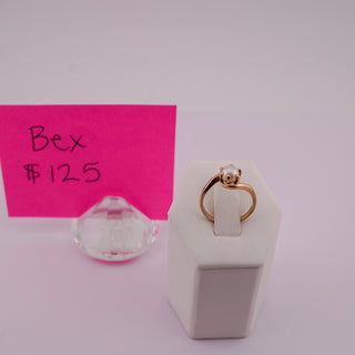 A gold ring with a clear round gemstone displayed on a white ring stand, next to a pink sticky note that reads 'Bex $125' on a light pink background.