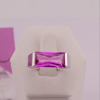 A rectangular magenta-pink gemstone ring with silver-toned metal borders, displayed on a white ring stand. A pink price card reading '$25 Marilyn' is visible in the background next to a clear glass sphere prop.