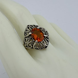 A silver filigree ring with a central oval orange gemstone, displayed on a cream-colored ring stand against a light gray background.