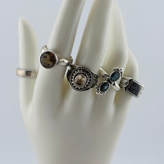 A set of four silver rings displayed on a white ceramic mannequin hand against a light gray background. The rings feature a smoky brown stone, a circular filigree design with a central clear stone, two small oval blue stones, and a rectangular silver inlay.
