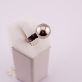 A silver ring with a polished spherical ball on a band, displayed on a white jewelry stand against a pale pink background.