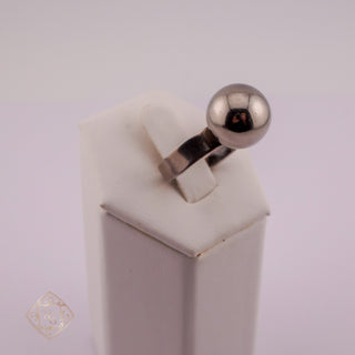 A silver ring with a polished spherical bead on a matching band, displayed on a white jewelry stand against a light gray background.