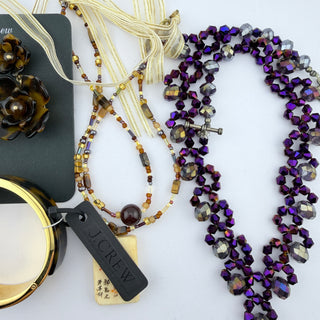 A J.Crew jewelry flat lay on a white background, featuring a multi-strand beaded necklace with purple and iridescent beads, a matching beaded tassel necklace, and a black brand tag.