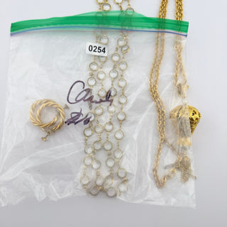 A clear plastic zip-top bag containing multiple jewelry items, including gold-tone chains, a beaded necklace with white beads, a twisted gold ring, and a gold pendant, photographed on a white background.