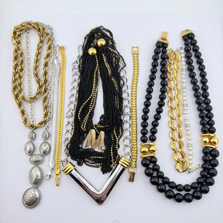A flat lay of multiple fashion necklaces arranged on a white background, including gold-tone and silver-tone chains, beaded strands, layered links, and black bead necklaces with gold and silver accents.