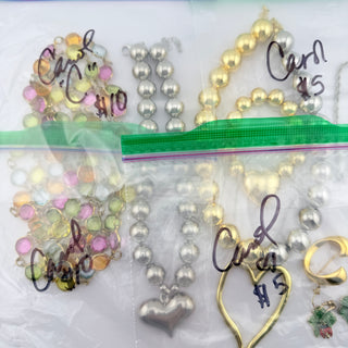 Several beaded necklaces and bracelets laid out on a clear plastic bag with handwritten price tags and labels. The jewelry includes pastel multicolor beads, silver beads with heart pendants, and gold beads with heart pendants.