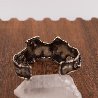 Reserved Grace artisan brutalist cuff bracelet in silver tone metal with organic textured edges on a wooden surface