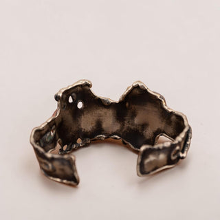 Reserved Grace artisan brutalist cuff bracelet in silver tone metal with organic textured edges on a wooden surface