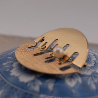 Reserved Grace gold tone abstract brooch with mixed finish and a single pearl accent on a blue floral surface